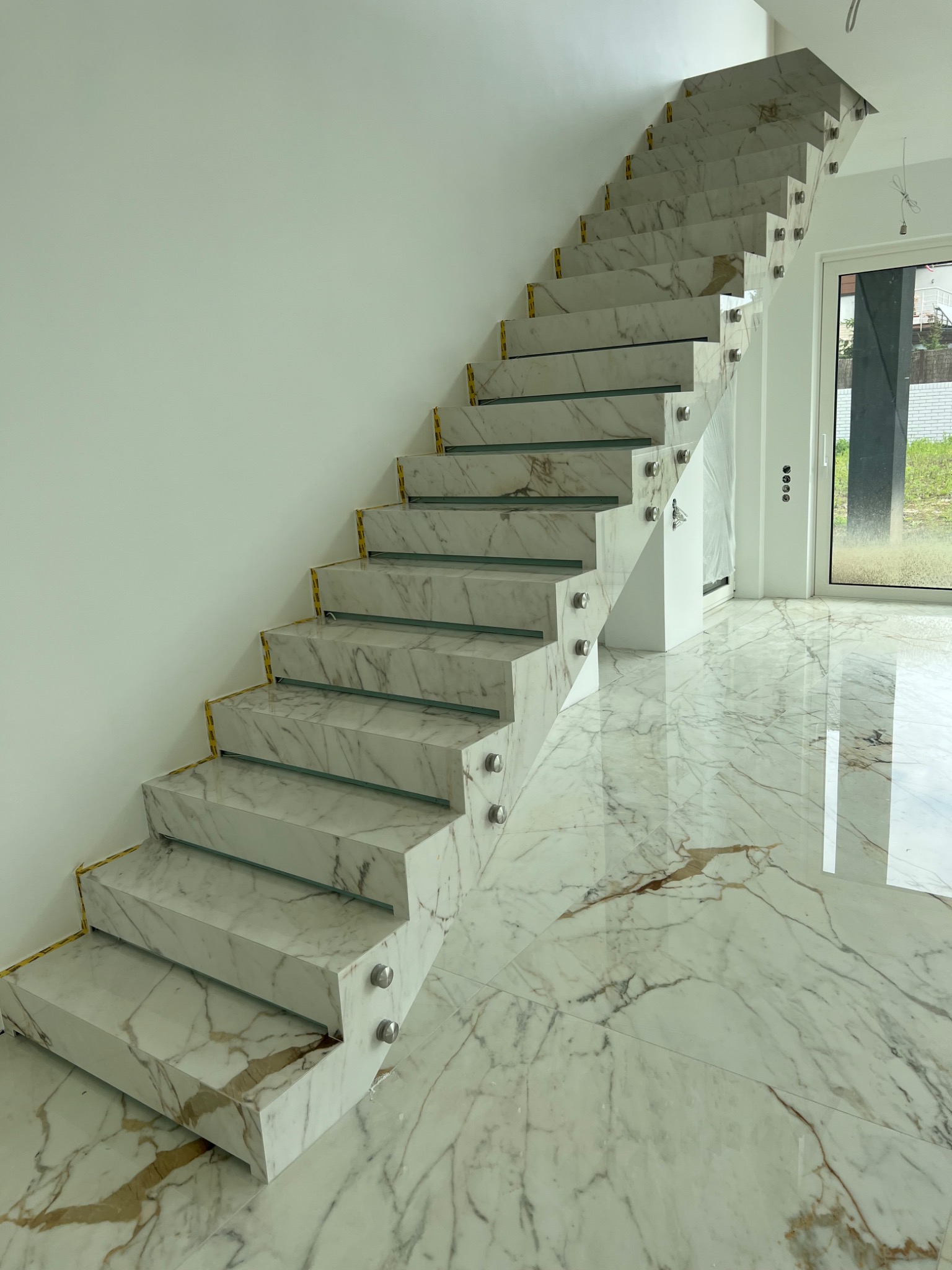 Marmurowe schody z balustradą ze szkła i metalu, widoczne z dołu. Podłoga wyłożona marmurem w tym samym wzorze. Nowoczesny design, jasne wnętrze.