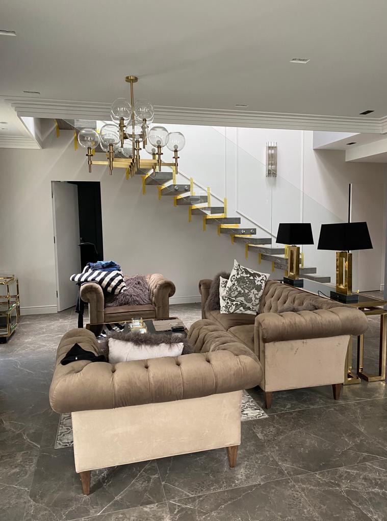 Salon z szarymi płytkami podłogowymi i schodami. Szklana balustrada, złote akcenty na schodach i lampach. Beżowe meble wypoczynkowe.