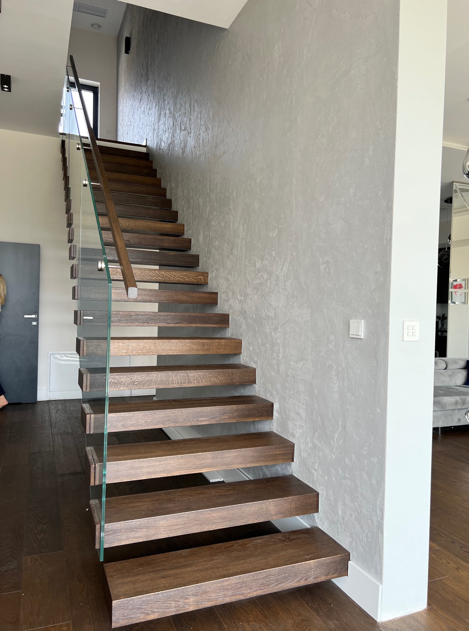 Nowoczesne, drewniane schody z szklaną balustradą w jasnym wnętrzu. Ściana wykończona dekoracyjnym tynkiem w odcieniach szarości. Widoczny fragment parkietu.