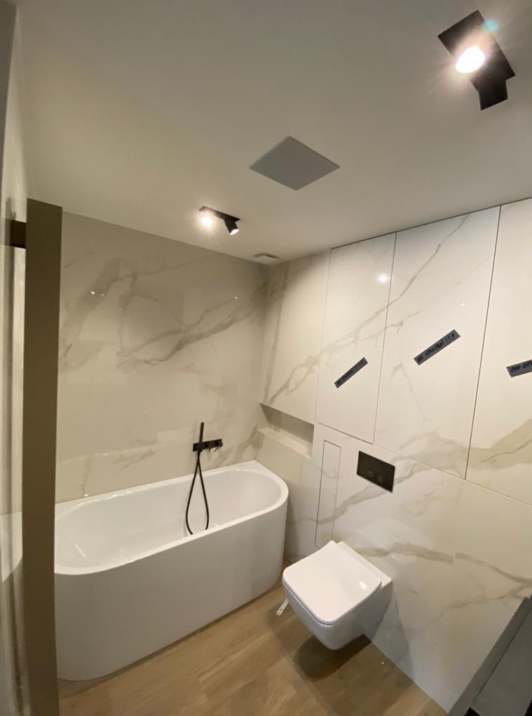 Nowoczesna łazienka po remoncie: biała wanna, toaleta, marmurowe płytki i oświetlenie punktowe. Widoczne czarne akcenty w postaci baterii i przycisku spłukiwania.