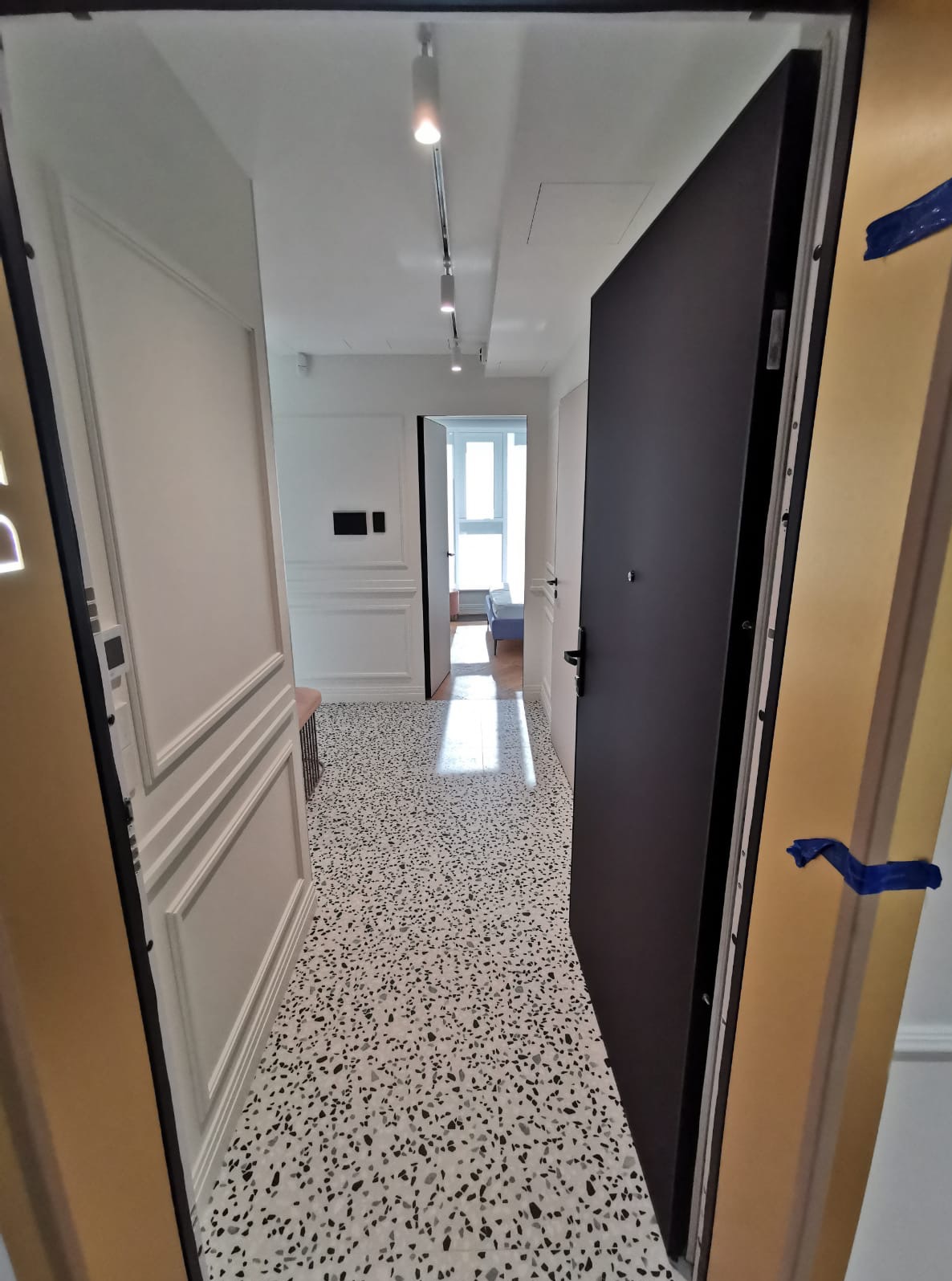 Nowoczesny przedpokój po remoncie: białe ściany z dekoracyjnymi panelami, lastryko na podłodze i czarne drzwi wejściowe. Widok z otwartych drzwi.