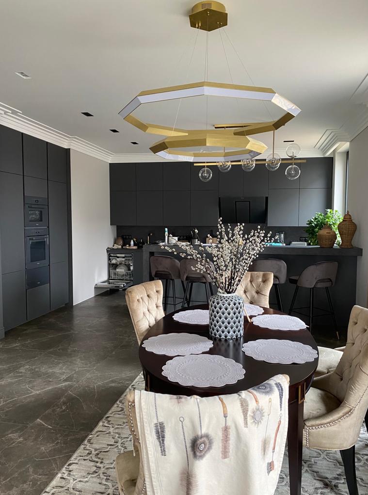 Nowoczesna kuchnia z ciemnymi szafkami, wyspą i okrągłym stołem z dekoracyjnym wazonem. Złoty, geometryczny żyrandol dodaje elegancji. Widoczne krzesła z beżową tapicerką.