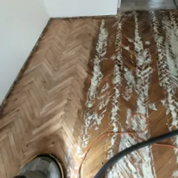 Częściowo wyszlifowany parkiet ułożony w jodełkę, z widocznymi śladami po maszynie do cyklinowania i resztkami starego lakieru. Fragment urządzenia do cyklinowania w rogu.