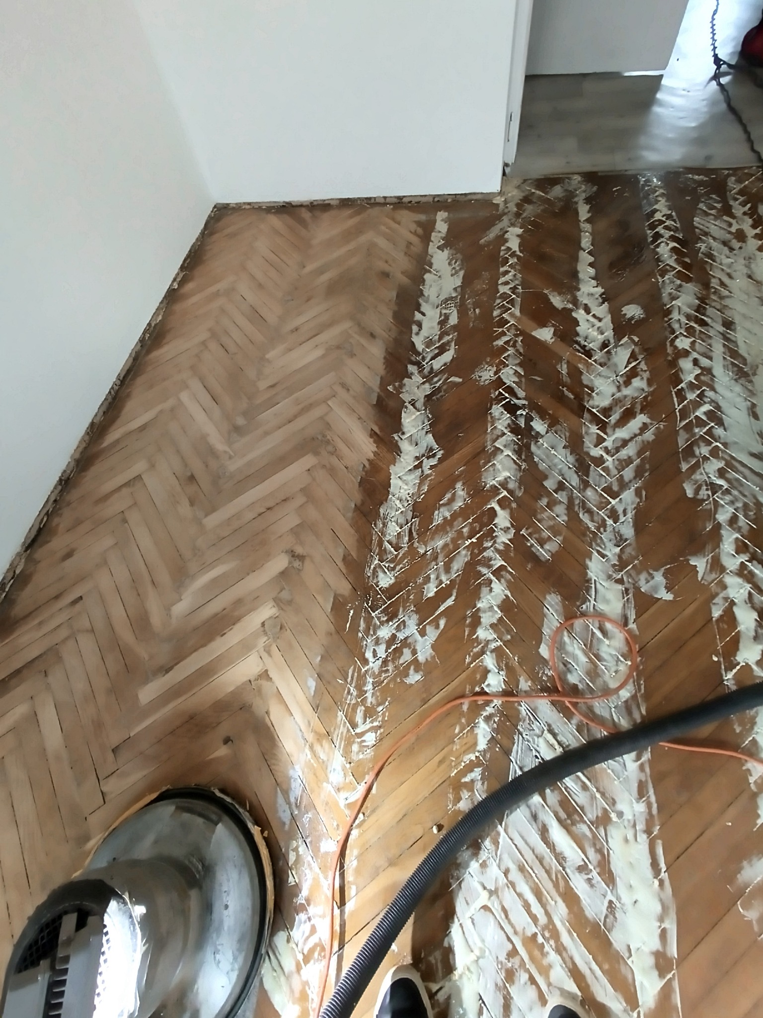 Częściowo wyszlifowany parkiet ułożony w jodełkę, z widocznymi śladami po maszynie do cyklinowania i resztkami starego lakieru. Fragment urządzenia do cyklinowania w rogu.