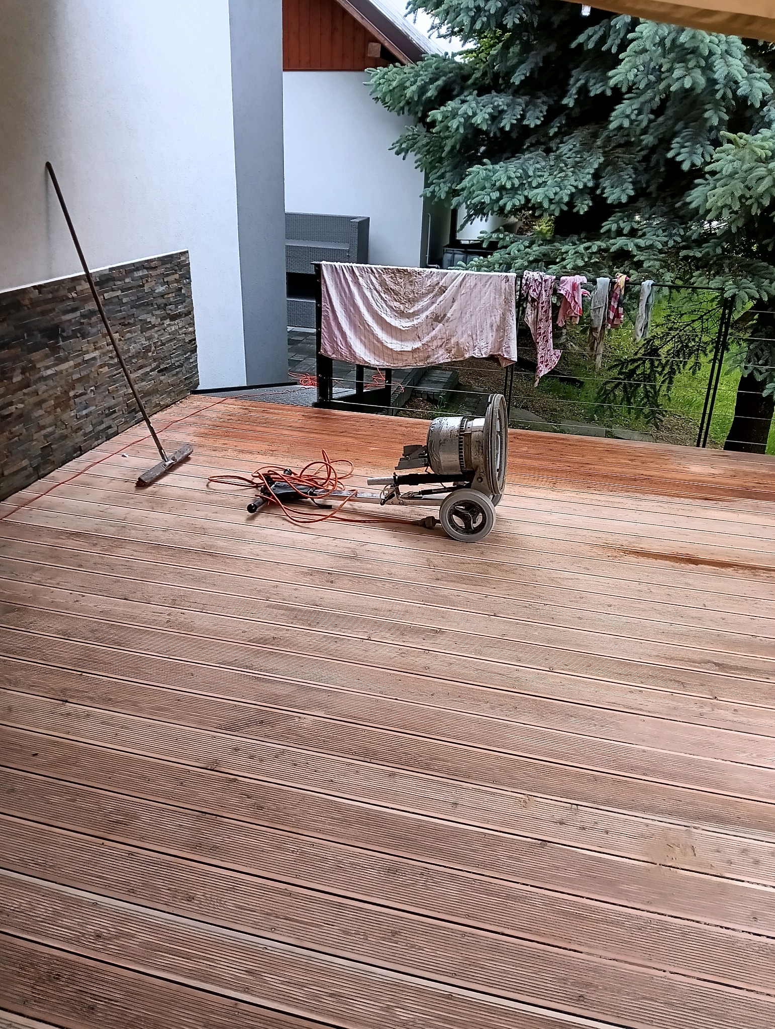 Cyklinowanie drewnianego tarasu zewnętrznego przy użyciu maszyny. Widoczna szczotka i pranie na suszarce w tle.