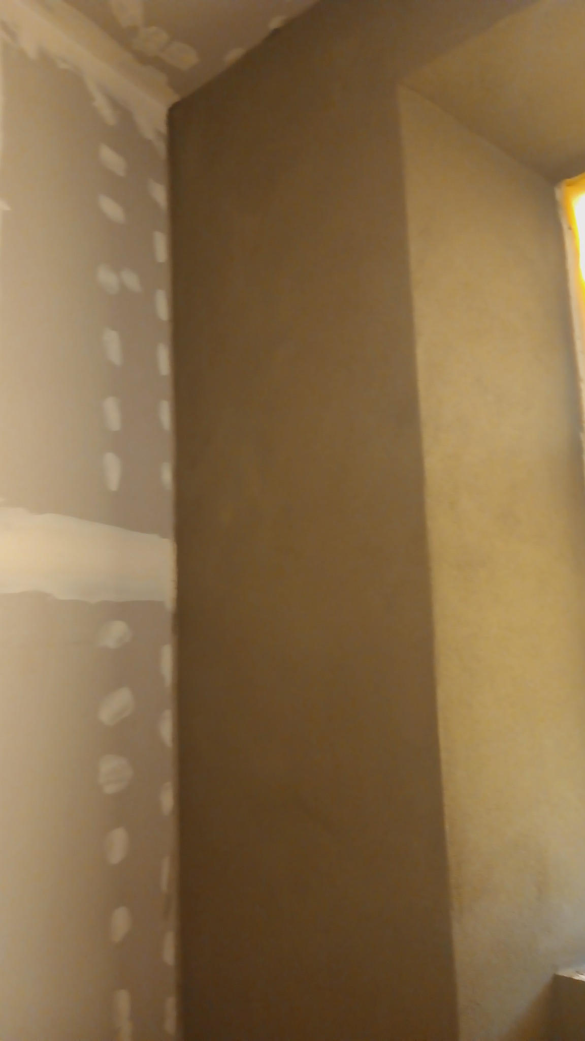 Ściana po nałożeniu gładzi w odcieniach beżu, widoczne fragmenty zaszpachlowanej ściany gipsowej w rogu pomieszczenia, przygotowanie do malowania.