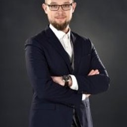 W&K GOS ACCOUNTING SPÓŁKA Z OGRANICZONĄ ODPOWIEDZIALNOŚCIĄ - Młody mężczyzna w okularach i granatowej marynarce, z założonymi rękami, na ciemnoszarym tle. Profesjonalny wizerunek.