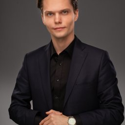 W&K GOS ACCOUNTING SPÓŁKA Z OGRANICZONĄ ODPOWIEDZIALNOŚCIĄ - Młody mężczyzna w granatowym garniturze i czarnej koszuli, z zegarkiem na ręku, pozuje na szarym tle. Profesjonalny wizerunek.