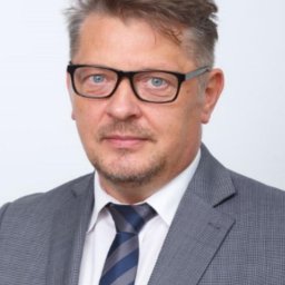 W&K GOS ACCOUNTING SPÓŁKA Z OGRANICZONĄ ODPOWIEDZIALNOŚCIĄ - Portret biznesowy mężczyzny w okularach, szarej marynarce i krawacie w paski na jasnym tle. Profesjonalny wygląd.