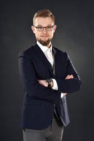 Młody mężczyzna w okularach i granatowej marynarce, z założonymi rękami, na ciemnoszarym tle. Profesjonalny wizerunek.