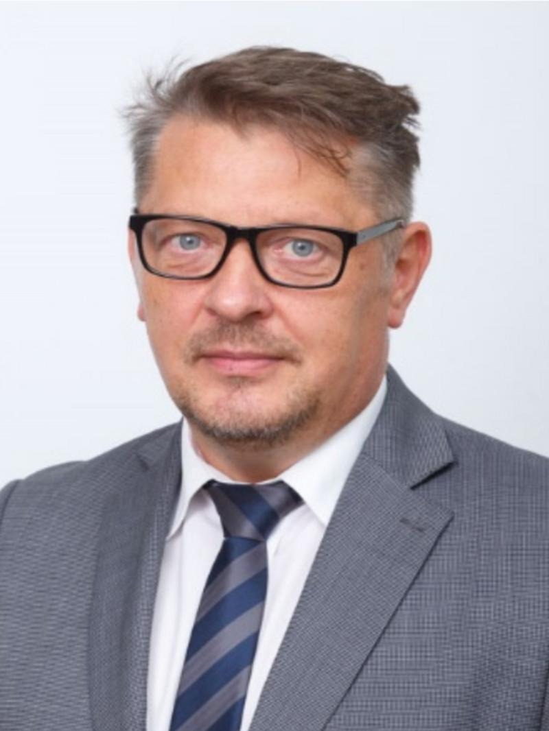 Portret biznesowy mężczyzny w okularach, szarej marynarce i krawacie w paski na jasnym tle. Profesjonalny wygląd.