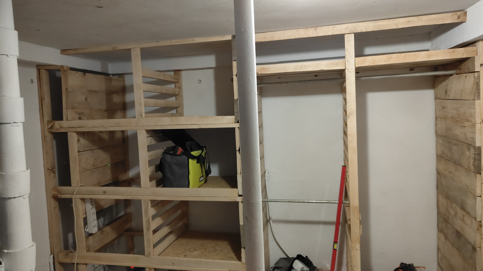 Drewniana konstrukcja garderoby w trakcie budowy. Widoczne półki, drążek na ubrania i torba narzędziowa. Ściany częściowo obłożone drewnem. Poziomica oparta o ścianę.