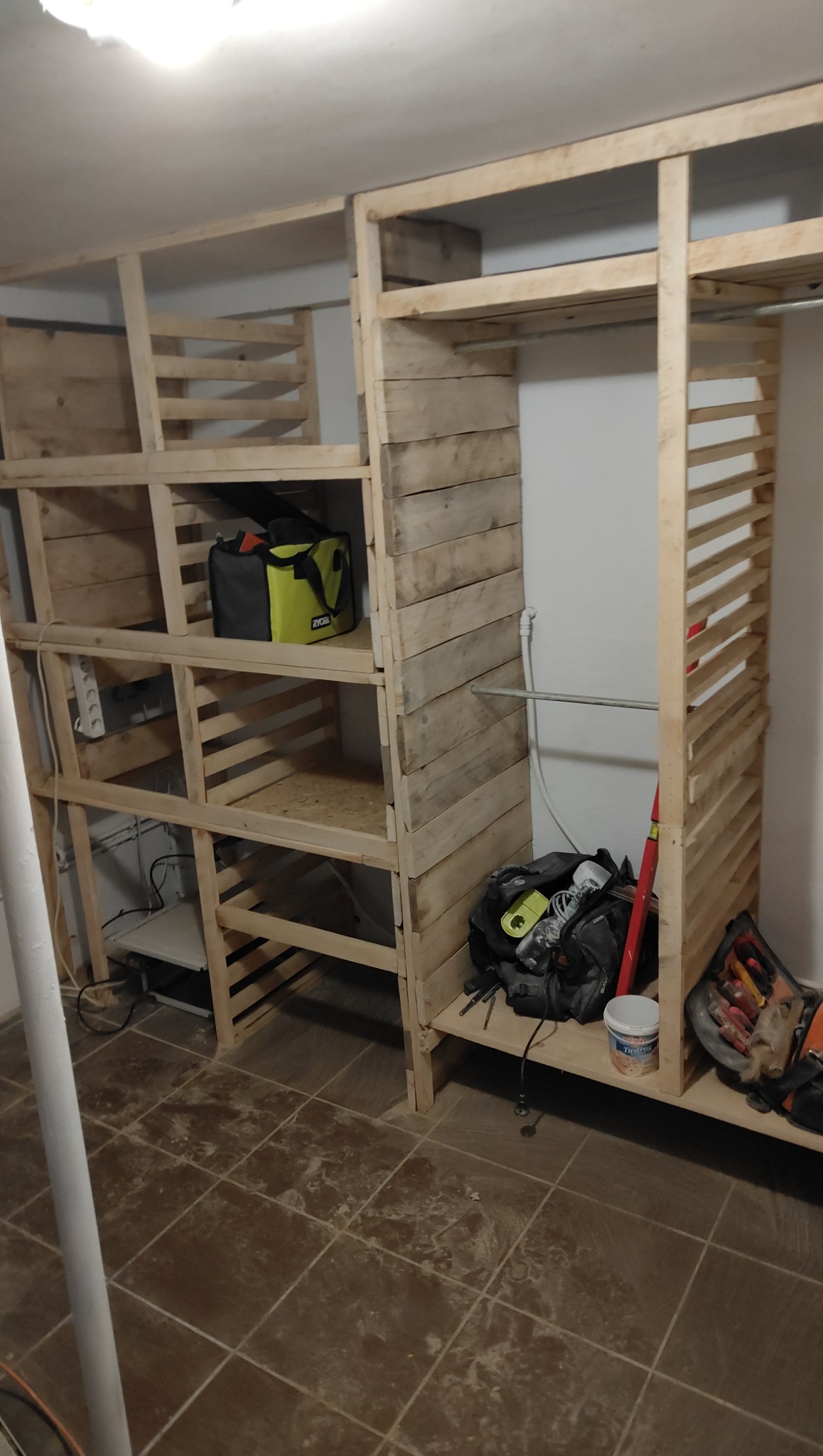 Konstrukcja garderoby z surowego drewna, z półkami i drążkiem, w trakcie montażu. Widoczne narzędzia i torba narzędziowa. Prosta forma, naturalne materiały.