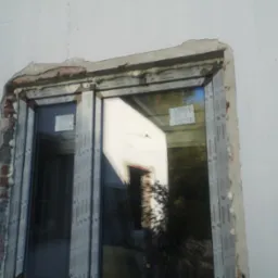 Montaż okna PCV marki Salamander w surowym otworze okiennym, widoczne pianowanie i fragment muru. W szybie odbija się budynek i zieleń.