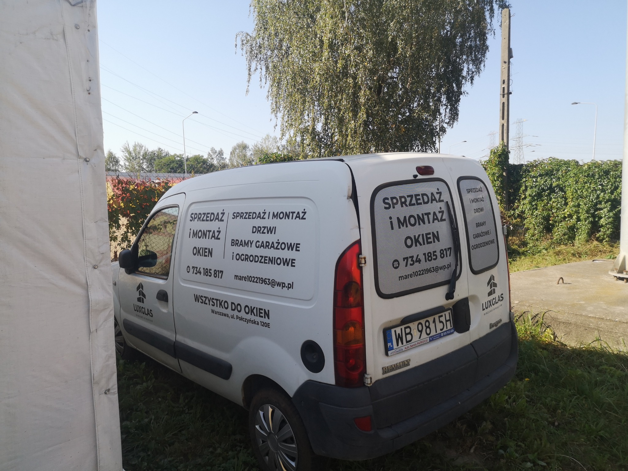 Biały samochód dostawczy z reklamą firmy Luxglas oferującej sprzedaż i montaż okien, drzwi, bram garażowych i ogrodzeniowych. Widoczne dane kontaktowe. Auto zaparkowane na zewnątrz.