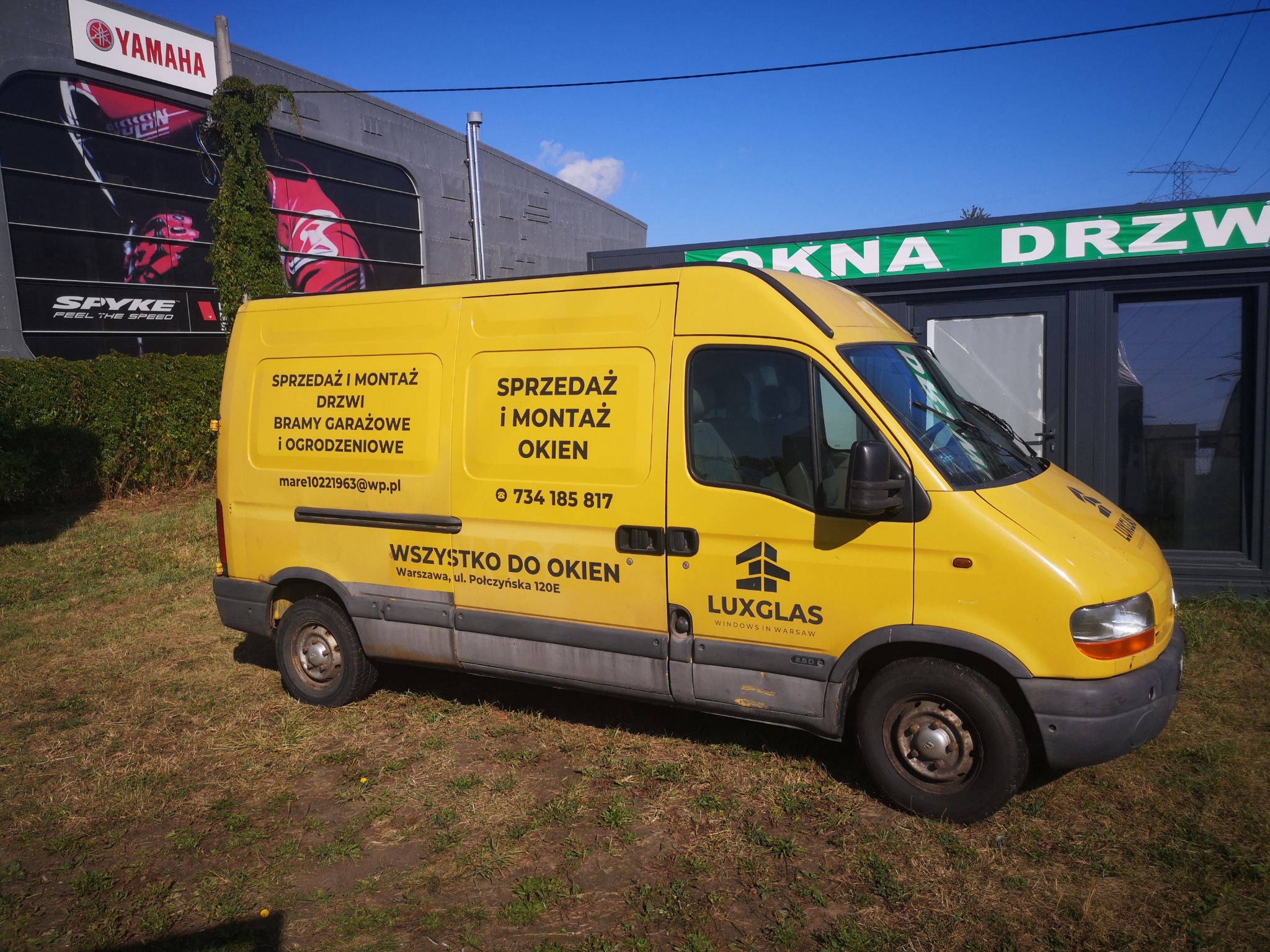 Żółty van firmy Luxglas z reklamą sprzedaży i montażu okien, drzwi i bram garażowych na tle budynku z reklamą Yamaha i ekspozycji okienno-drzwiowej.