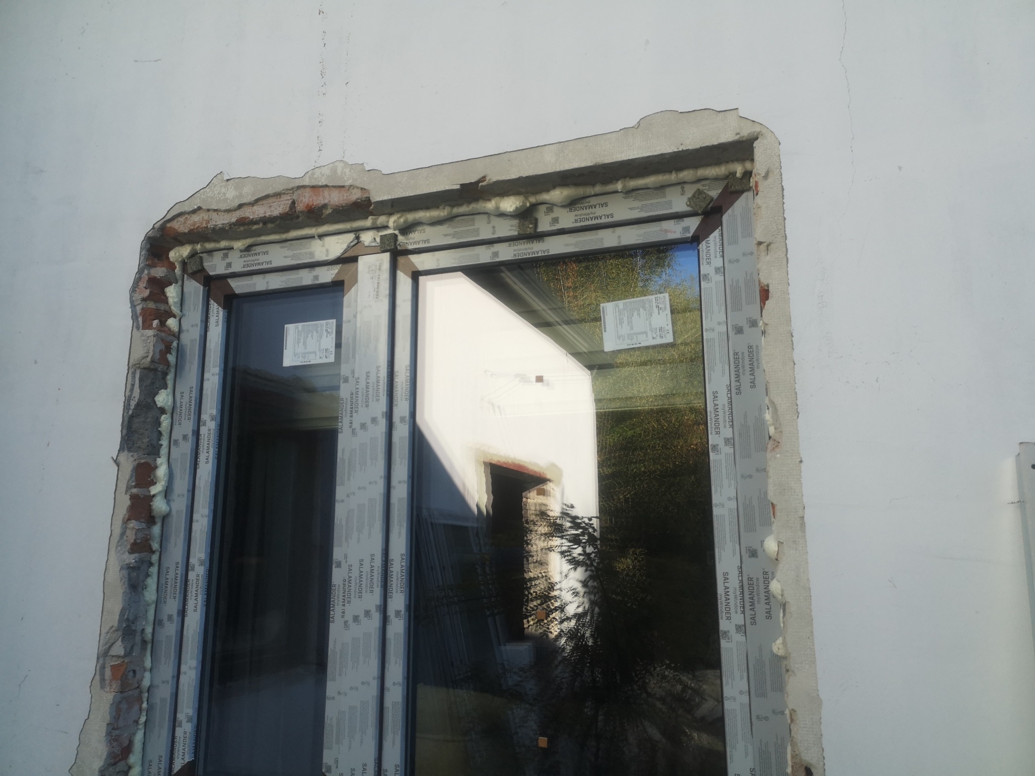Montaż okna PCV marki Salamander w surowym otworze okiennym, widoczne pianowanie i fragment muru. W szybie odbija się budynek i zieleń.