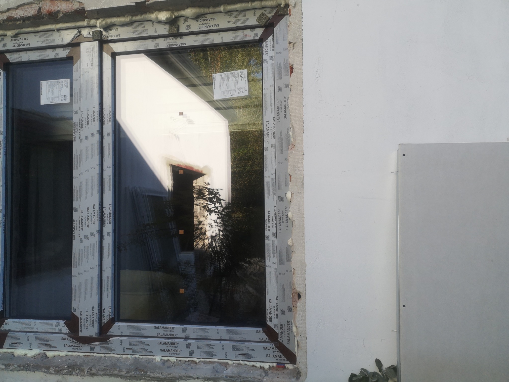 Dwukomorowe okno z folią ochronną marki Salamander, widoczne pianowanie montażowe. W szybie odbija się zieleń drzew. Częściowo otynkowana ściana.