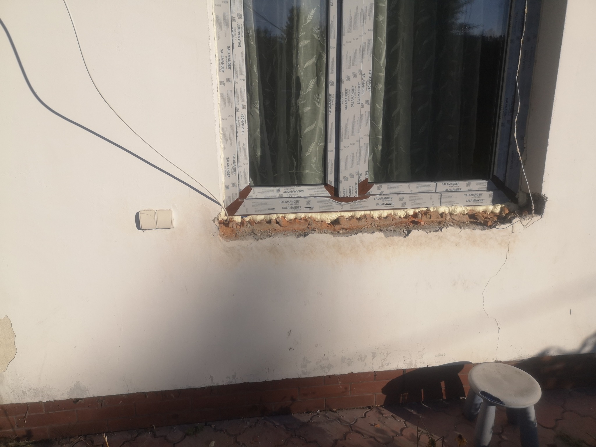 Okna PCV Salamander po montażu, widoczne pianka montażowa i uszkodzenia ściany. Ujęcie z zewnątrz, z dołu, widać fragment chodnika i stołek.