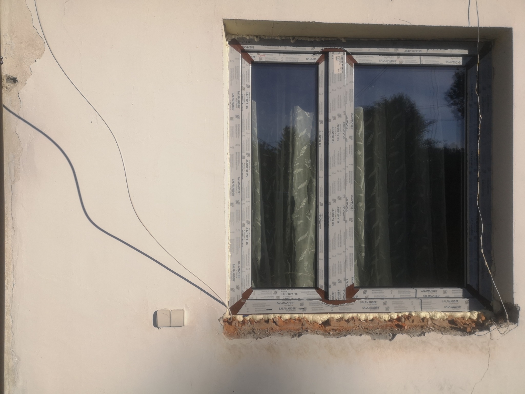 Szare okno PCV Salamander, zamontowane w ścianie z widocznymi śladami pianki montażowej i tynku. Widoczne przewody elektryczne i refleksy zieleni w szybie.