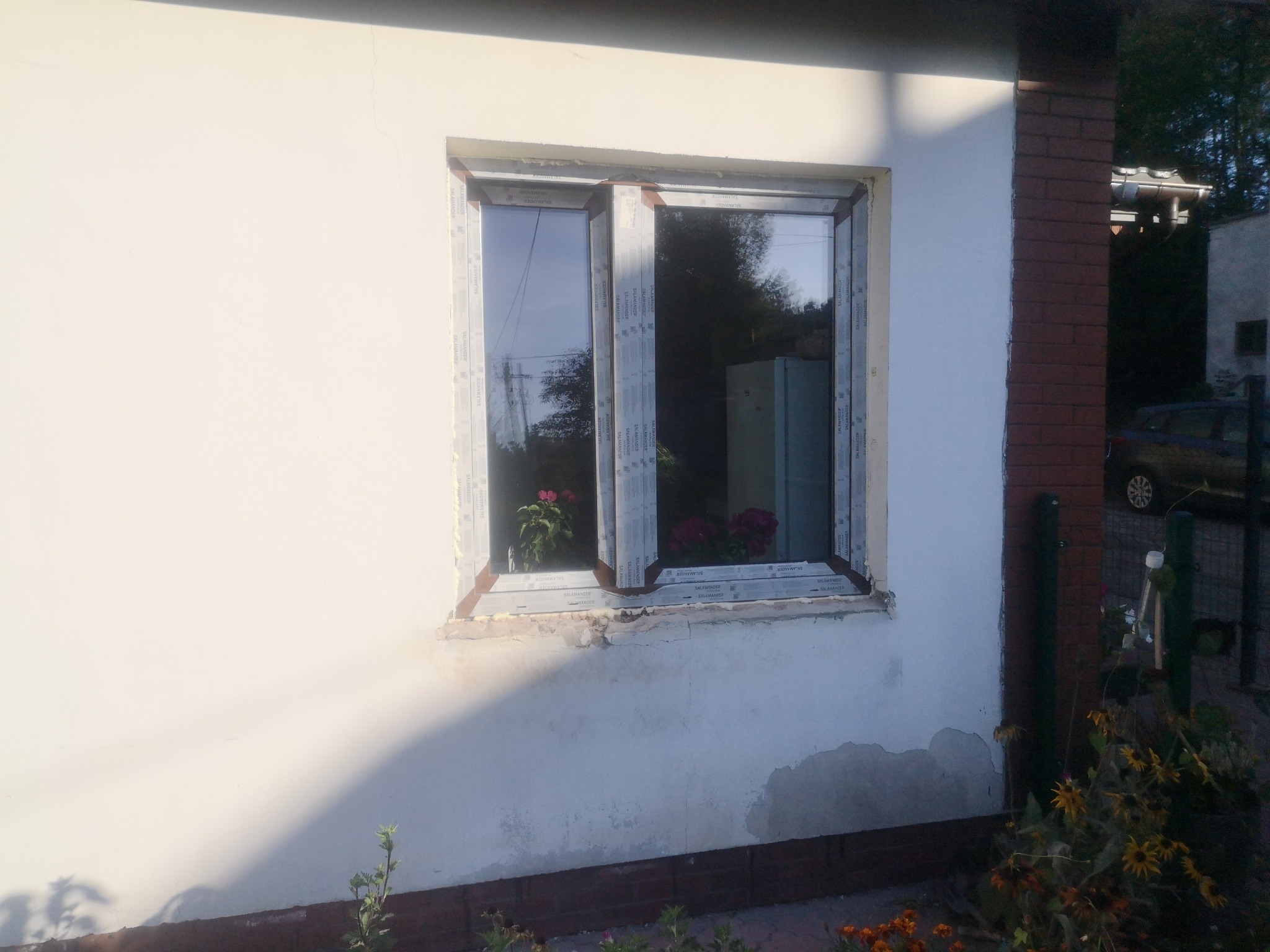 Białe okno PCV Salamander w otynkowanej ścianie domu, z kwiatami na parapecie. Widoczne ślady po starym oknie i niezakończony montaż. Częściowo oświetlone słońcem.