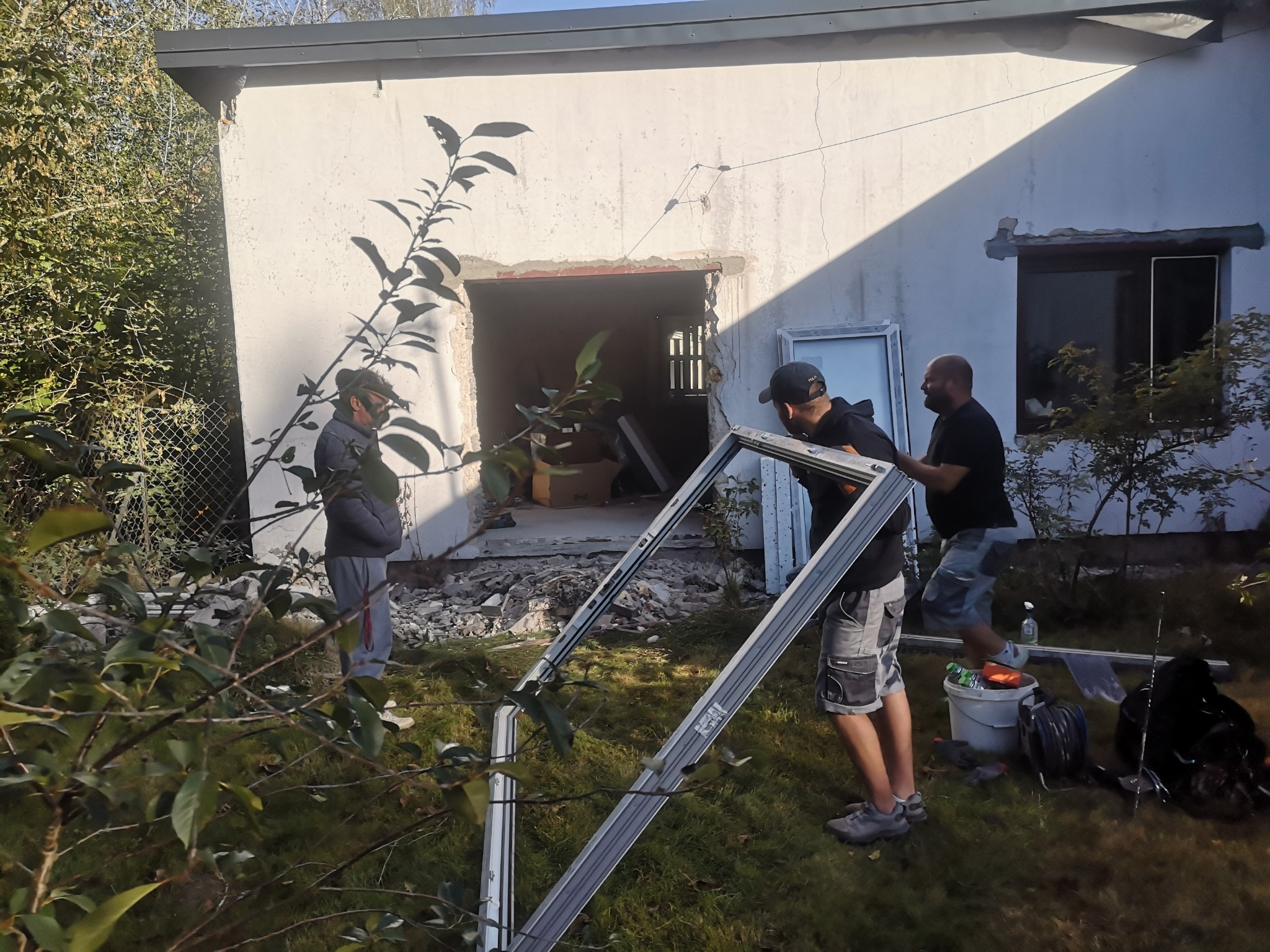 Dwóch monterów instaluje ramę okienną w budynku z gruzem. Widoczny starszy mężczyzna obserwujący pracę. Na trawie leżą narzędzia i akcesoria.