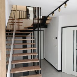 FusionWW - Nowoczesne schody z drewnianymi stopniami i czarną metalową konstrukcją, uzupełnione szklaną balustradą, w minimalistycznym wnętrzu z oświetleniem punktowym.