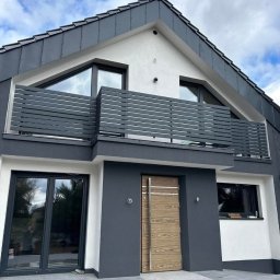 FusionWW - Nowoczesny dom z szarą, horyzontalną balustradą balkonową i drewnianymi drzwiami wejściowymi. Elewacja w odcieniach szarości i bieli. Minimalistyczny design.