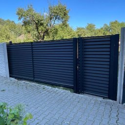 FusionWW - Nowoczesna, pozioma brama wjazdowa z czarnych, metalowych paneli, z widocznym fragmentem betonowego słupka i kostki brukowej. Brama na tle zieleni i błękitnego nieba.