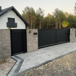 Nowoczesna, pozioma brama wjazdowa z czarnych paneli, wkomponowana w szare, kamienne filary i jasną kostkę brukową, z domem w tle i zielenią drzew. Elegancki design.