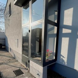 FusionWW - Szklana zabudowa balkonu z czarną ramą, widok z zewnątrz. Wewnątrz kwiaty w doniczce. Szary budynek w tle, słoneczny dzień.