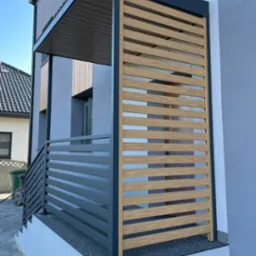 Nowoczesna zabudowa balkonu z drewnianymi lamelami i szarą metalową konstrukcją, widoczna z zewnątrz budynku. Minimalistyczny design i stonowana kolorystyka.