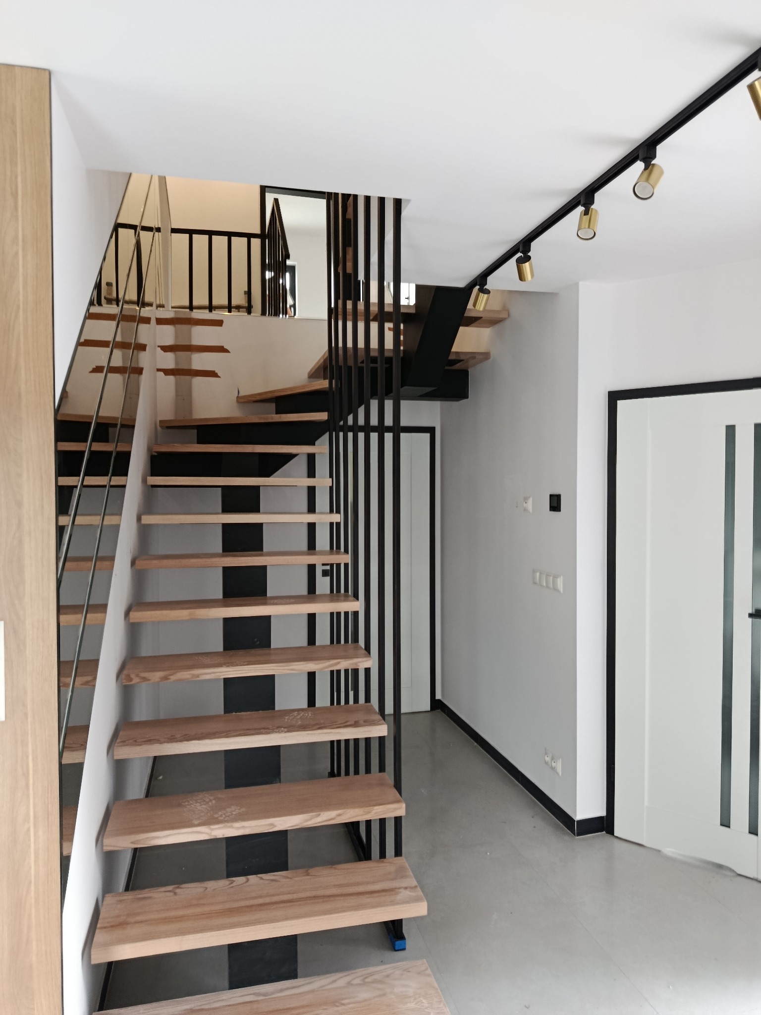 Nowoczesne schody z drewnianymi stopniami i czarną metalową konstrukcją, uzupełnione szklaną balustradą, w minimalistycznym wnętrzu z oświetleniem punktowym.