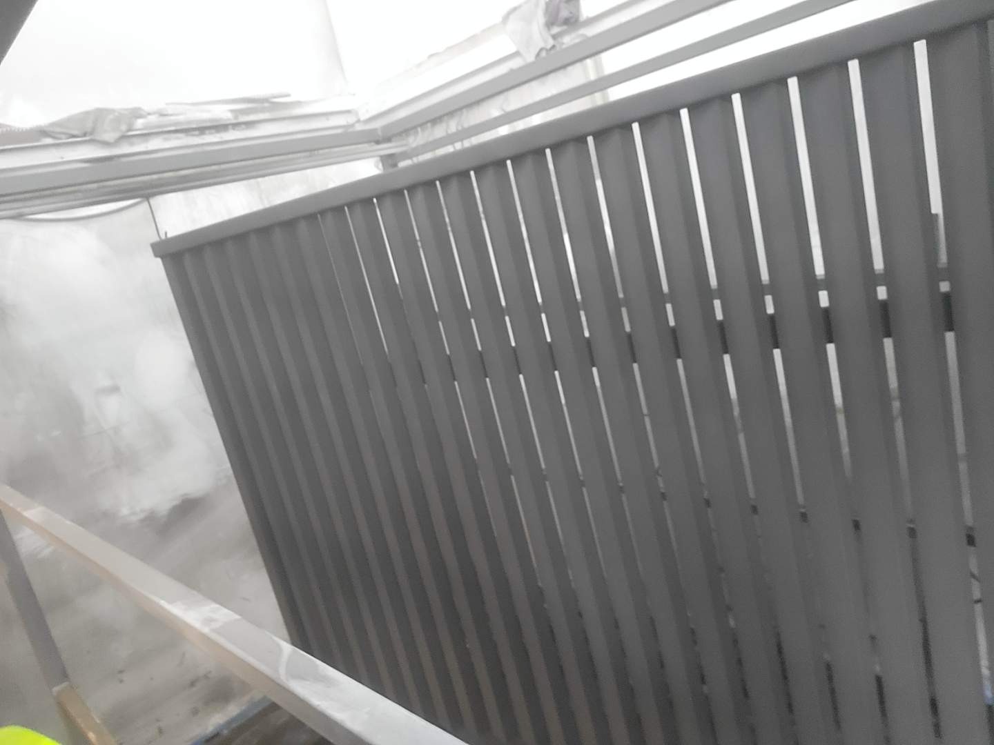 Szara, pionowa zabudowa balkonu z metalowych profili, widoczna z bliska, z elementami konstrukcyjnymi w tle. Minimalistyczny design.