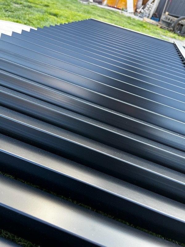 Czarne, metalowe lamele ogrodzeniowe ułożone równolegle na trawie, widok z góry pod kątem. Elementy przygotowane do montażu.