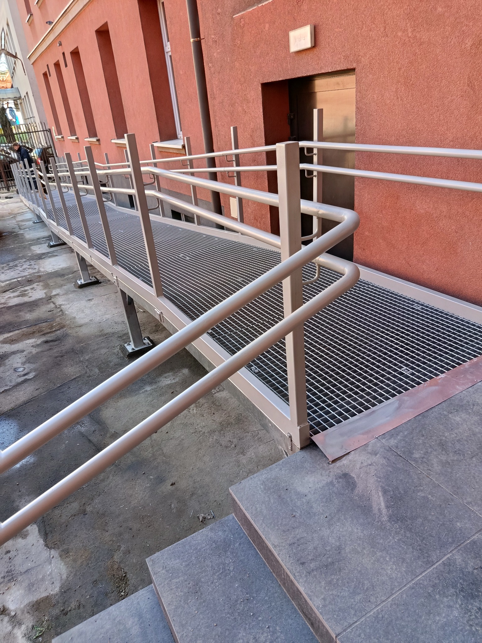 Metalowa rampa z poręczami dla niepełnosprawnych w Ogrodzieńcu. Konstrukcja z kraty pomostowej, prowadząca do drzwi budynku. Widoczne schody i elewacja.