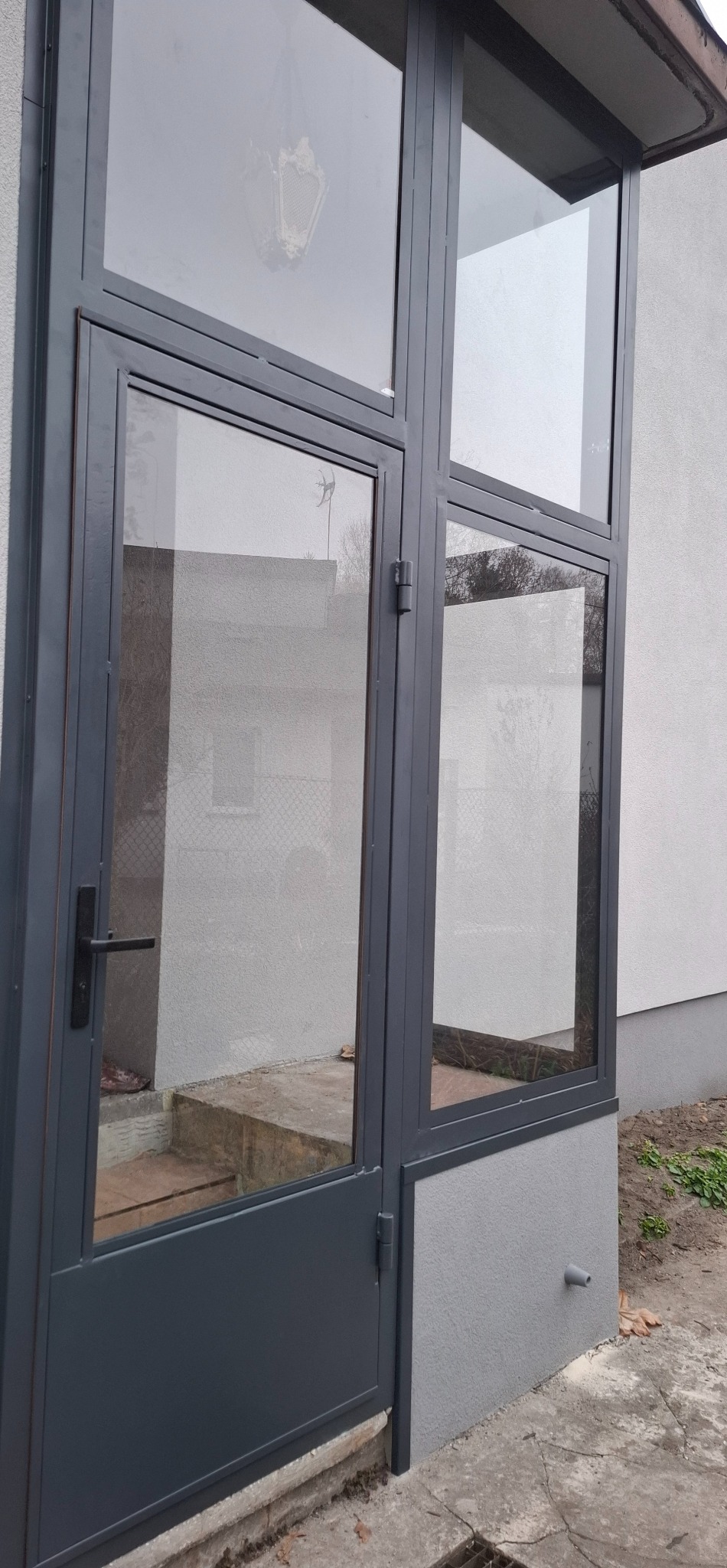 Szara metalowa konstrukcja z szybami, prawdopodobnie wejście do budynku lub zabudowa balkonu. Widoczne schody wejściowe i fragment elewacji budynku.