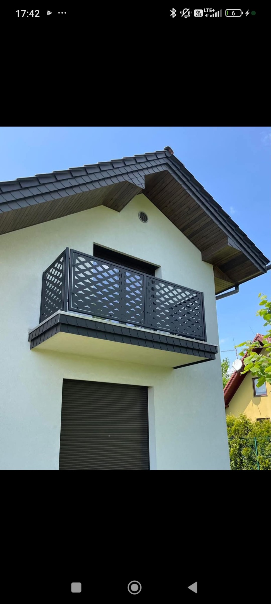 Balkon z nowoczesną, ażurową balustradą w kolorze grafitowym, dopasowaną do dachu z dachówki bitumicznej. Elewacja budynku biała, okno z roletą zewnętrzną.