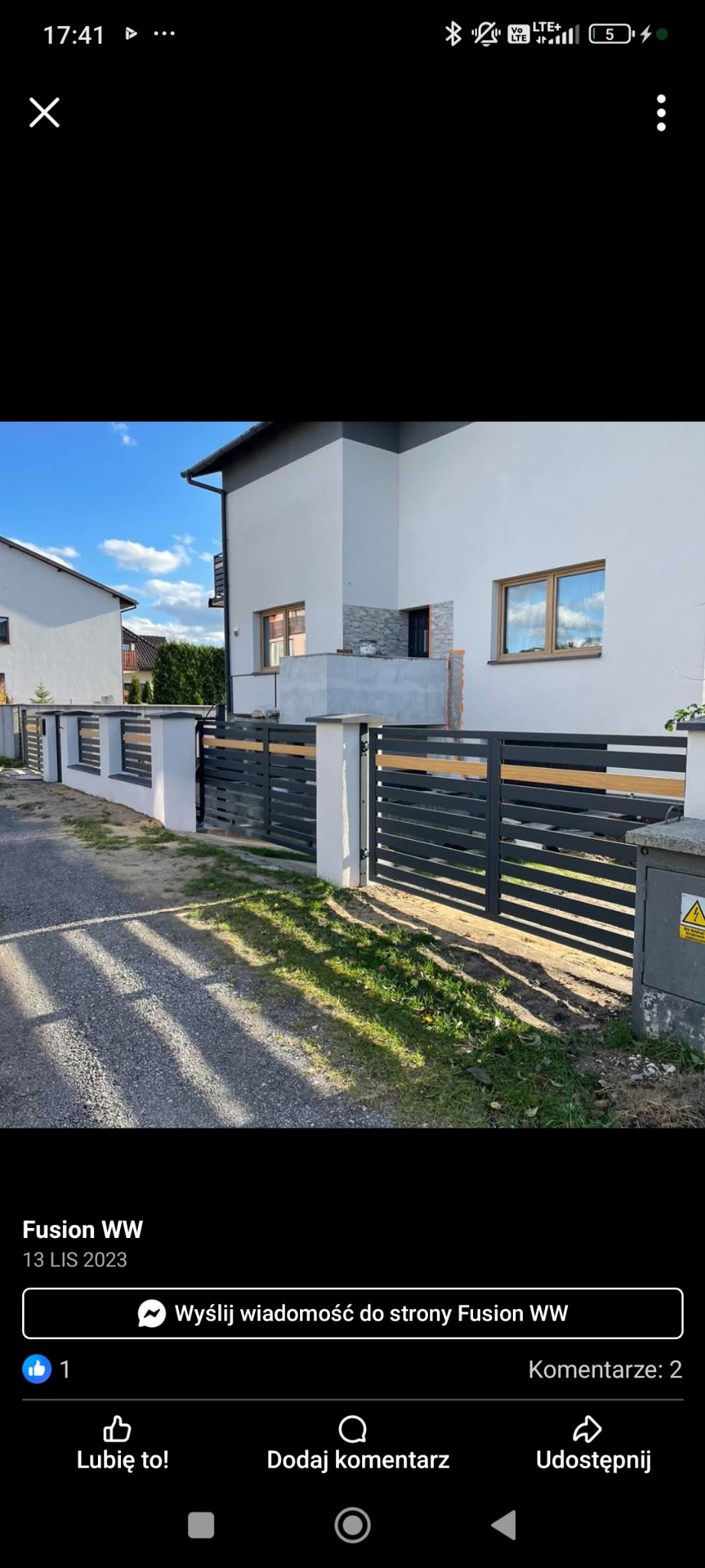 Nowoczesne ogrodzenie metalowe z drewnianymi akcentami przed domem z białego tynku. Brama wjazdowa zamknięta, widoczna skrzynka elektryczna.