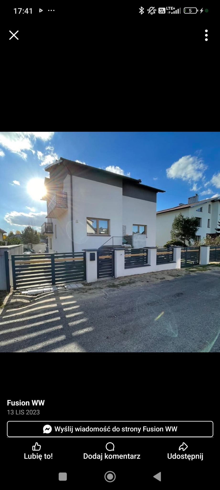 Nowoczesne ogrodzenie panelowe z poziomymi, ciemnymi przęsłami i jasnymi słupkami na tle dwukondygnacyjnego domu w słoneczny dzień. Brama przesuwna częściowo otwarta.