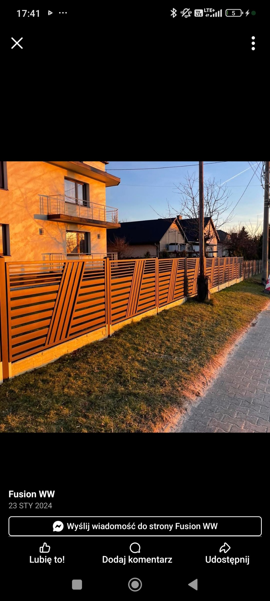 Nowoczesne ogrodzenie poziome z elementami ukośnymi, wykonane z brązowych desek na betonowej podmurówce, w tle dom jednorodzinny.