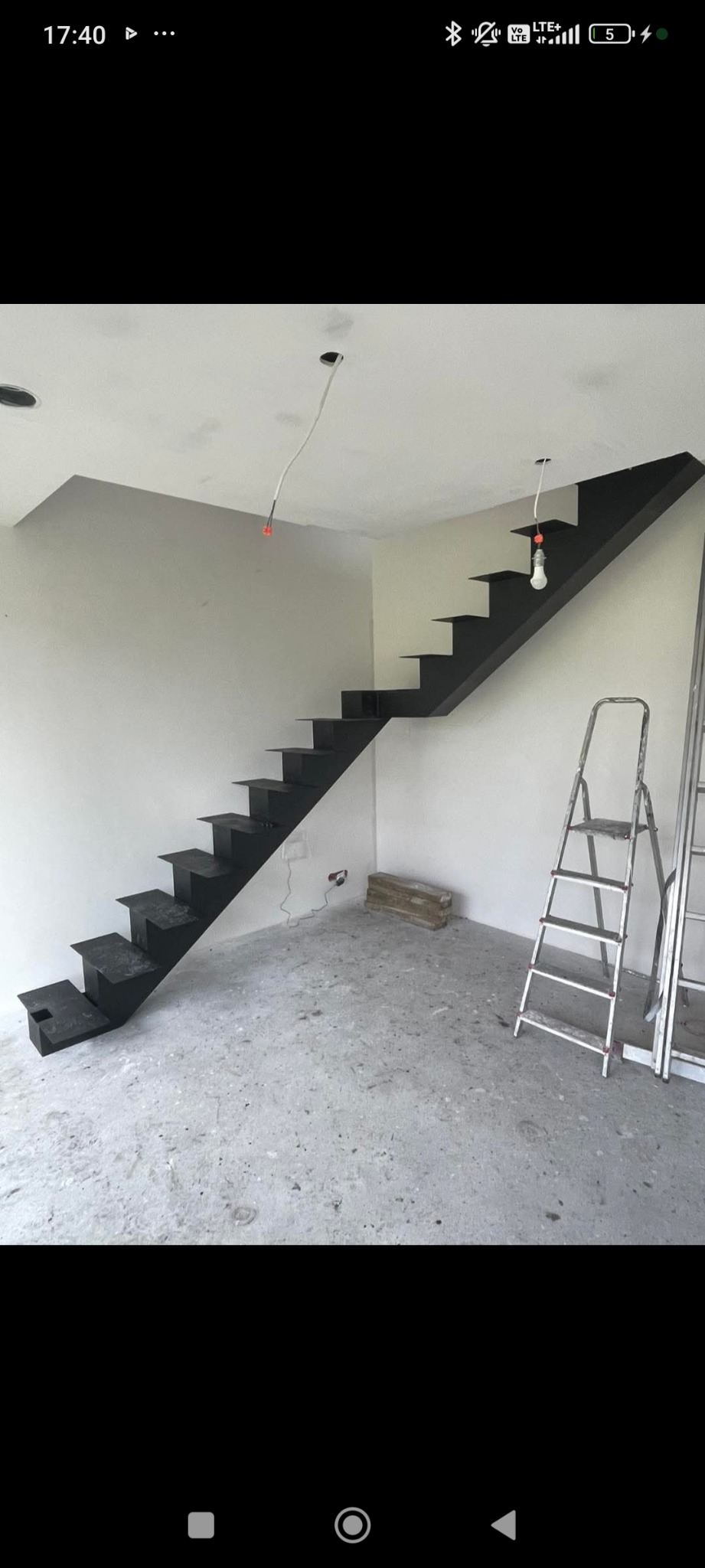 Nowoczesne, czarne schody metalowe wspornikowe w surowym wnętrzu, z widoczną drabiną i przewodami elektrycznymi zwisającymi z sufitu. Minimalistyczny design.