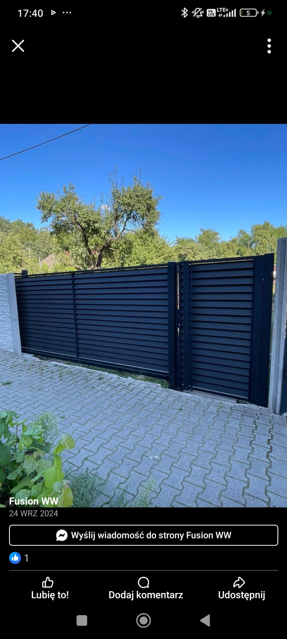 Nowoczesna, pozioma brama wjazdowa z czarnych, metalowych paneli, z widocznym fragmentem betonowego słupka i kostki brukowej. Brama na tle zieleni i błękitnego nieba.