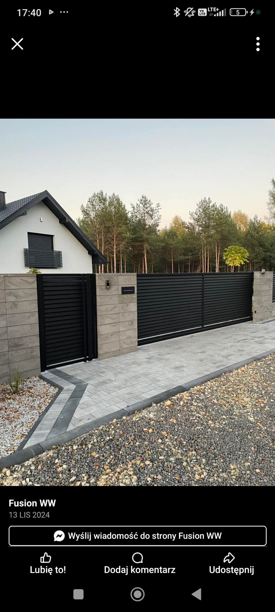 Nowoczesna, pozioma brama wjazdowa z czarnych paneli, wkomponowana w szare, kamienne filary i jasną kostkę brukową, z domem w tle i zielenią drzew. Elegancki design.