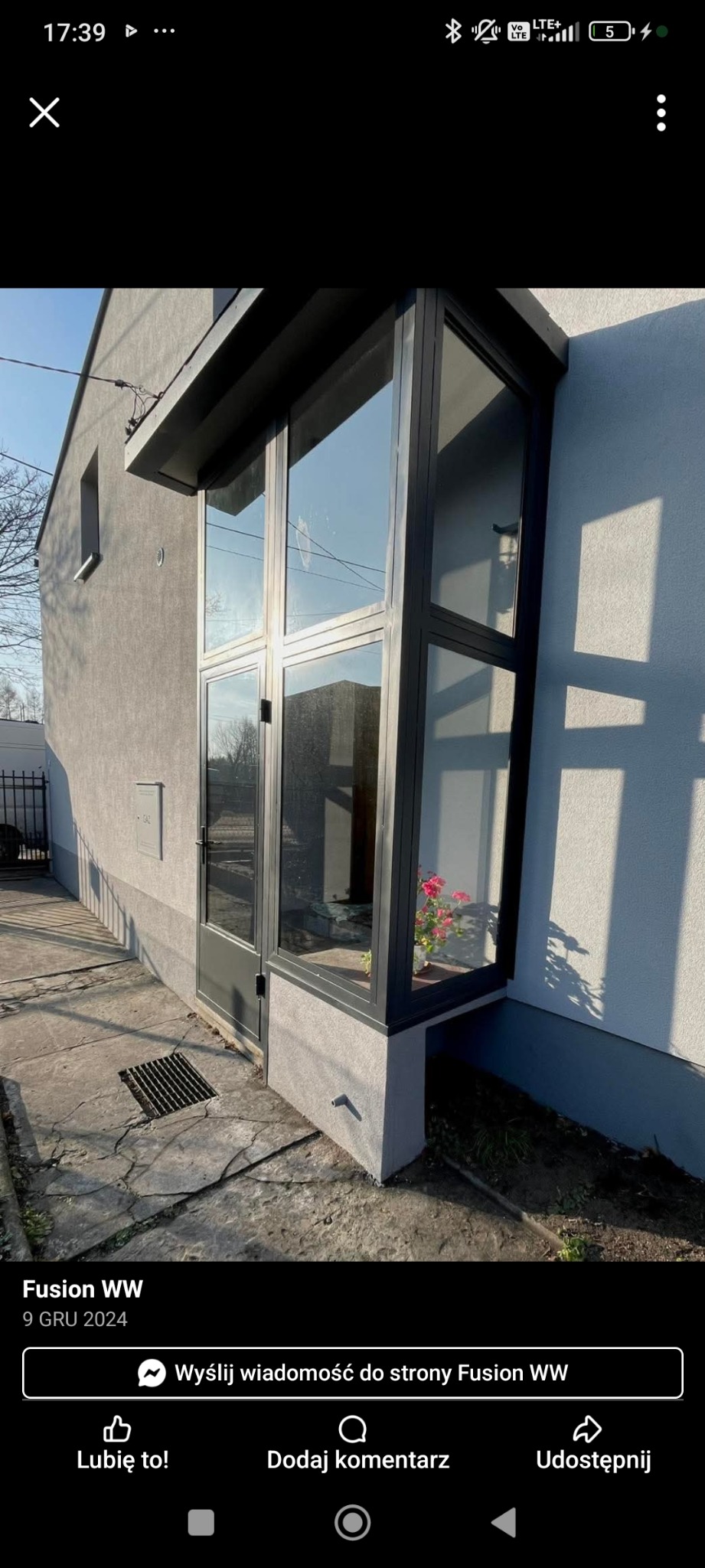 Szklana zabudowa balkonu z czarną ramą, widok z zewnątrz. Wewnątrz kwiaty w doniczce. Szary budynek w tle, słoneczny dzień.