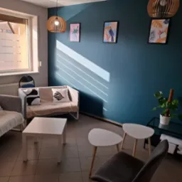 Jasne, minimalistyczne wnętrze gabinetu z turkusową ścianą, sofami z poduszkami w geometryczne wzory i drewnianymi lampami. Słońce wpada przez okno.