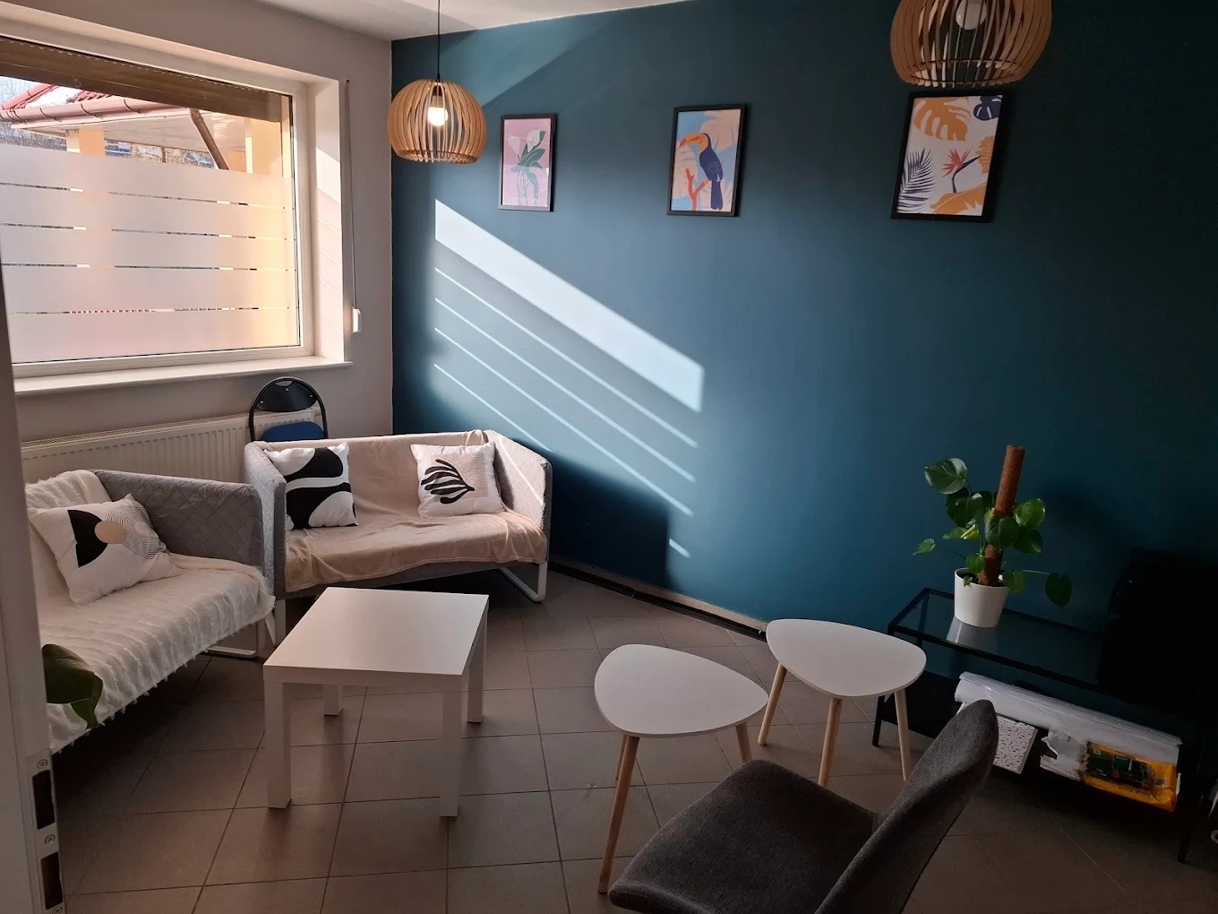 Jasne wnętrze gabinetu z sofami, stolikami i dekoracjami ściennymi w tonacji błękitu i bieli. Przytulna aranżacja z rośliną doniczkową i lampą wiszącą.