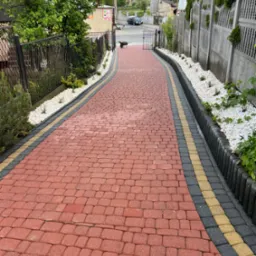 Czerwona kostka brukowa z obrzeżami w odcieniach szarości i żółci, otoczona białymi kamieniami i zielenią, prowadząca w dół w kierunku bramy i ulicy.