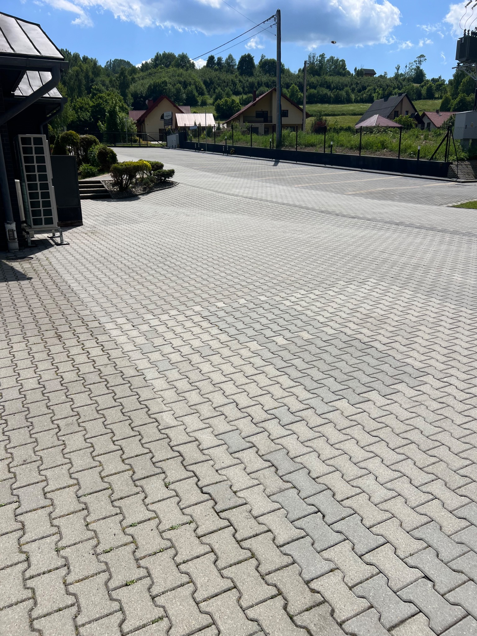 Rozległy plac wyłożony kostką brukową w kształcie litery H, otoczony zielenią i domami. Widok na czysty, jasny bruk w słoneczny dzień.