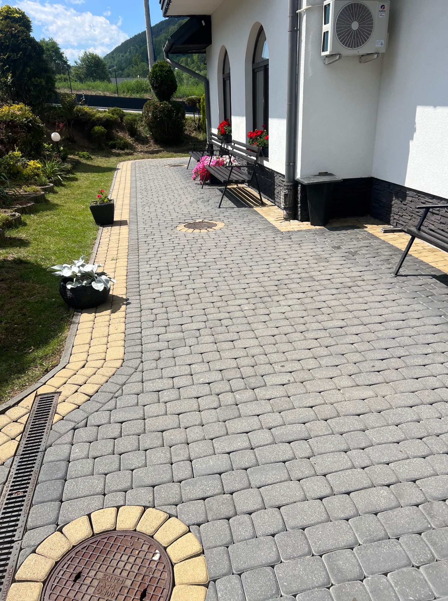 Ułożona szara kostka brukowa z żółtymi obrzeżami, widoczny fragment budynku z białymi ścianami, donice z kwiatami i zieleń ogrodu w tle.